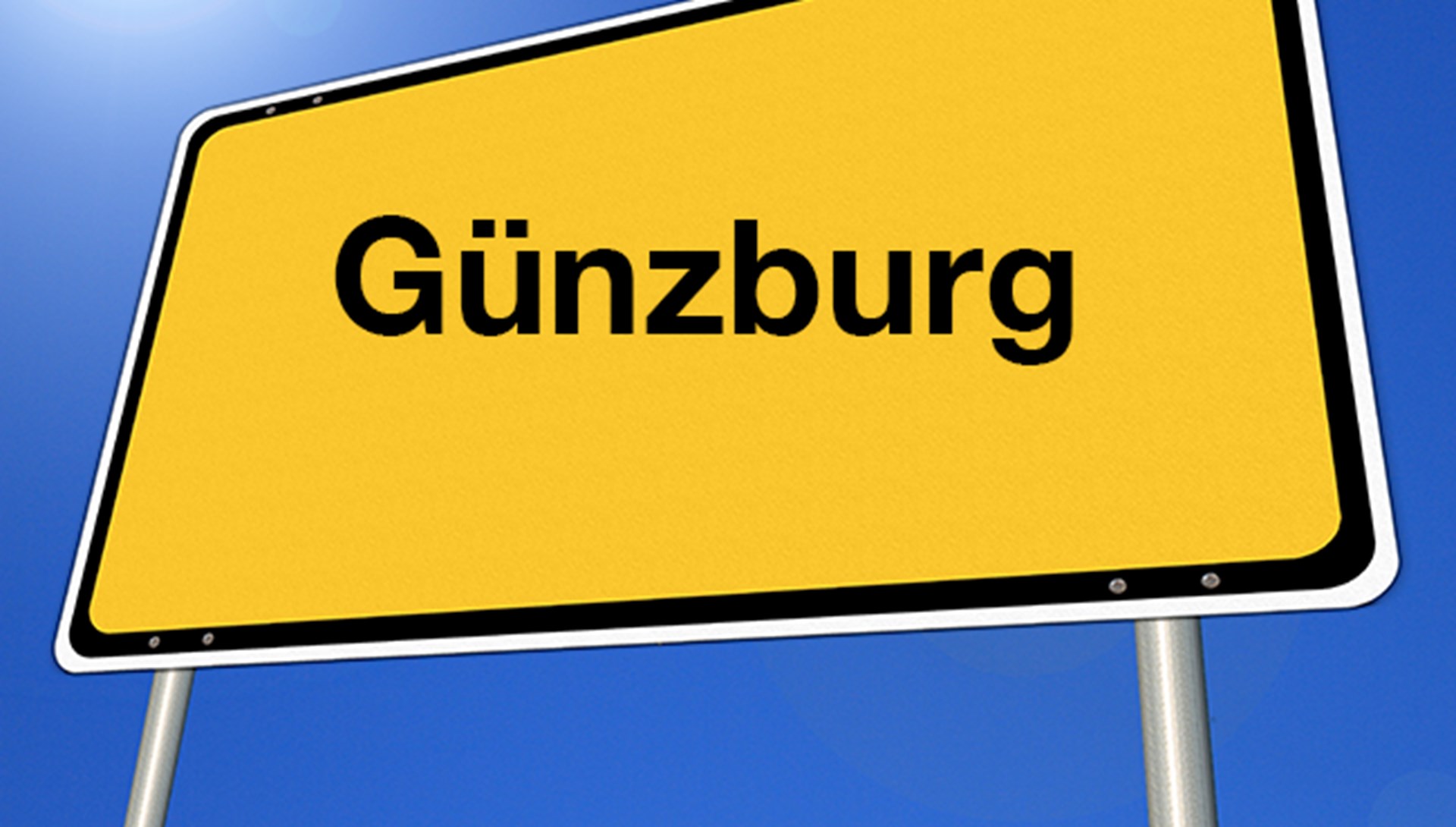 Das Foto zeigt ein schräg verzerrtes gelbes Ortsschild mit der Aufschrift Günzburg. 
